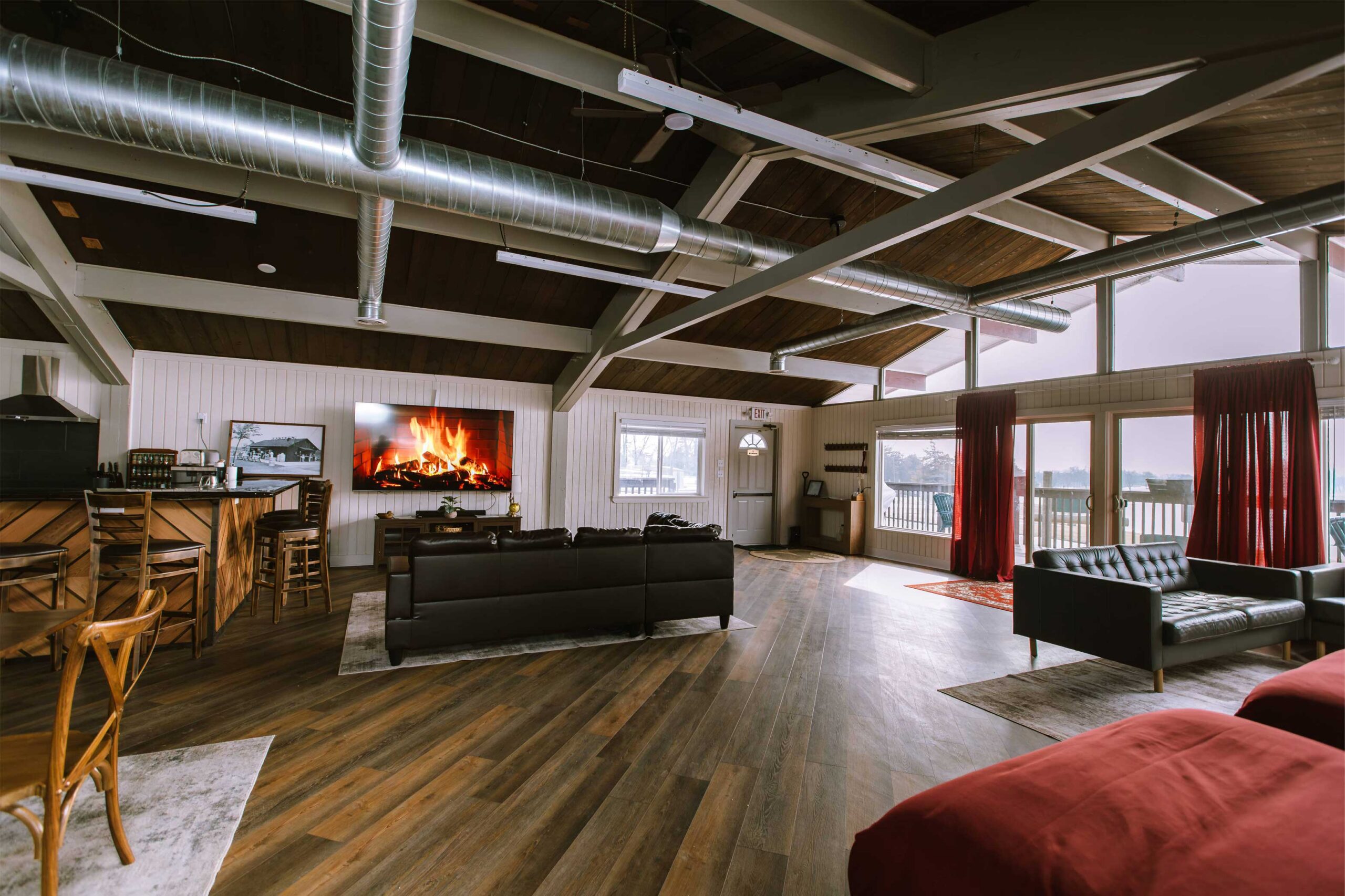 Cozy living room with a fireplace on TV, wooden flooring, and large windows with red curtains.
