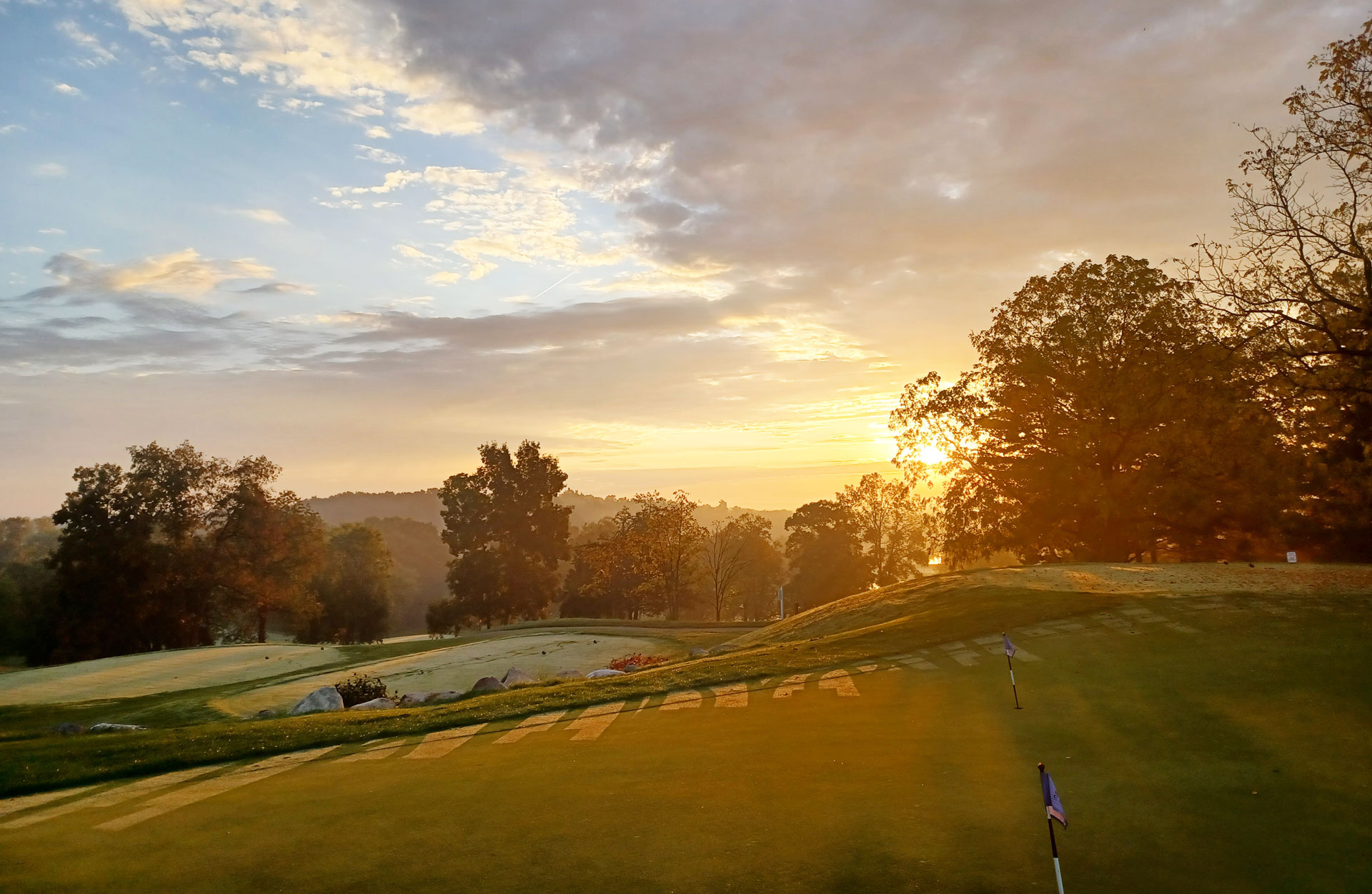 Sunrise over a golf course with trees and rolling hills, casting a warm glow on the landscape.