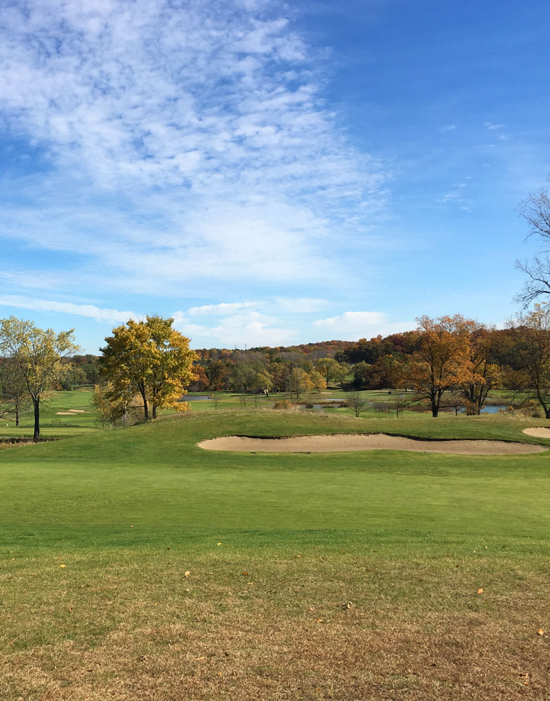 Scenic view of a golf course with autumn trees and a blue sky, featuring sand traps and lush green grass.