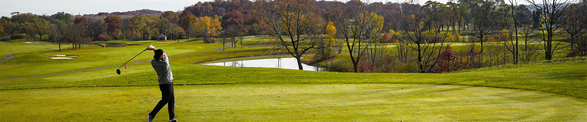 A golfer swings a club on a lush green golf course with trees and a pond in the background during autumn.