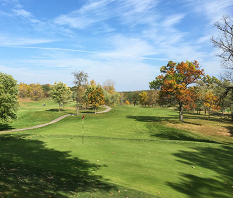 A scenic view of a golf course with trees displaying autumn colors and a flag on the green.
