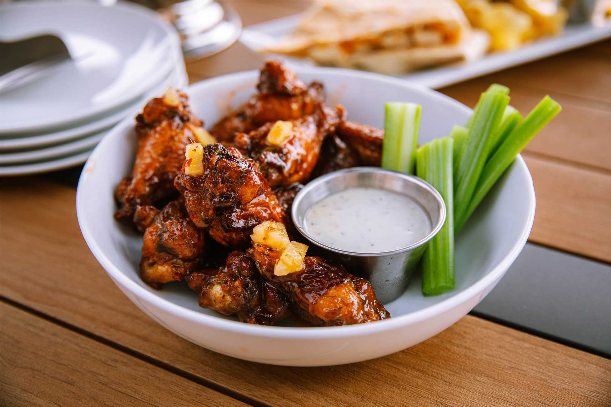 A bowl of glazed chicken wings with celery sticks and a small cup of dipping sauce on a wooden table.