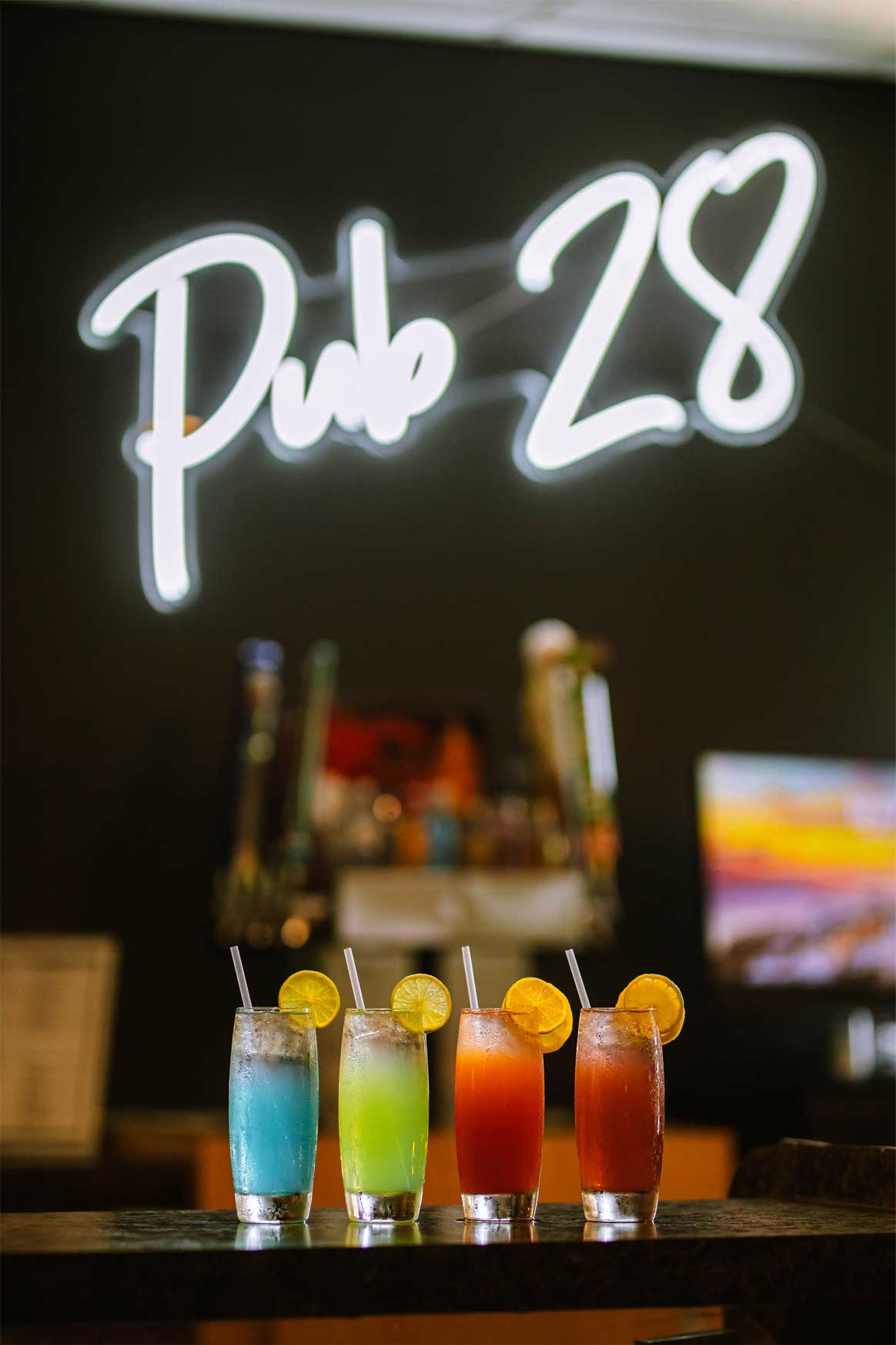 Four colorful cocktails with lemon slices on a bar, with a neon sign reading 'Pub 28' in the background.