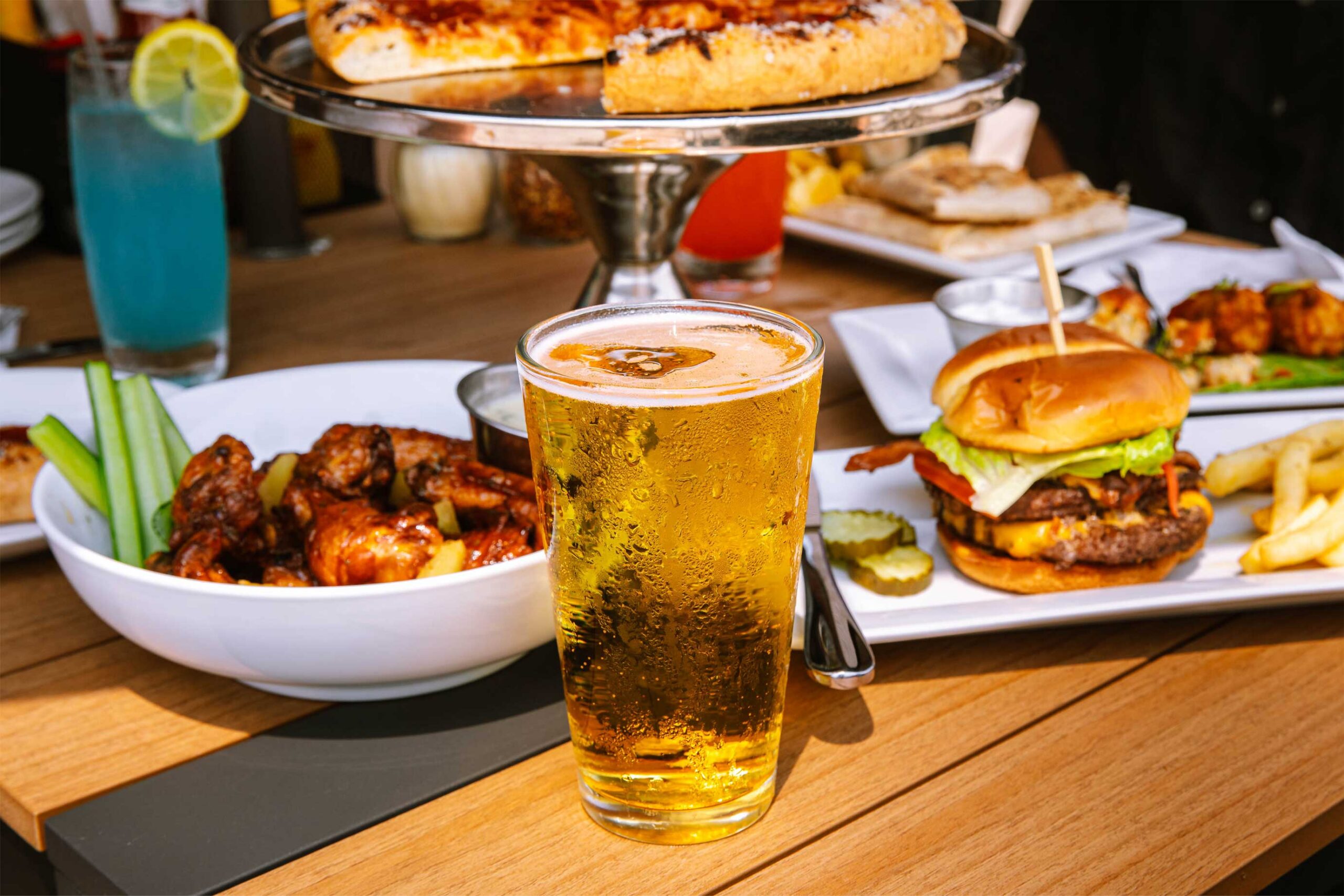 A glass of beer in front of a plate of chicken wings, a burger, and a side of fries on a wooden table.