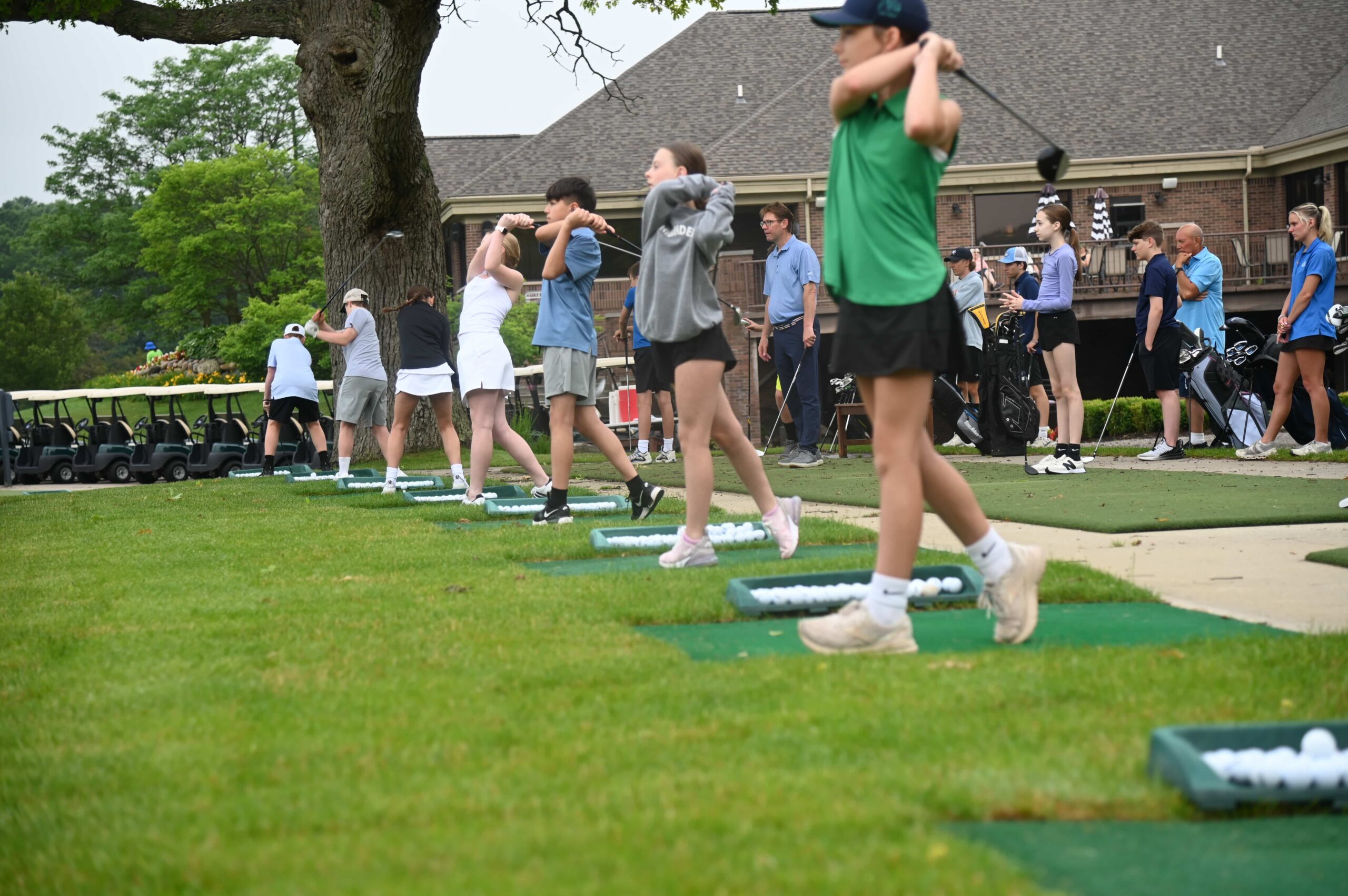 Young golfers practicing swings at a driving range, with golf carts and instructors in the background.