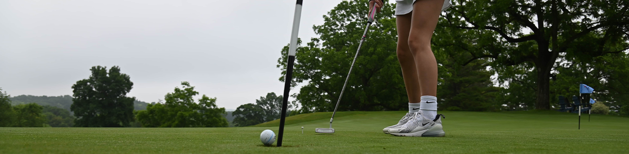 A golfer prepares to putt on a green, with a golf ball near the hole and trees in the background.